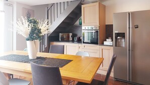 Una cocina moderna con horno empotrado, microondas y refrigerador. Hay una mesa de comedor con sillas y un florero. Una escalera conduce a un nivel superior.