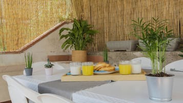 Een ontbijtarrangement met een tafel, stoelen, een dienblad met croissants, sap en melk, en potplanten.