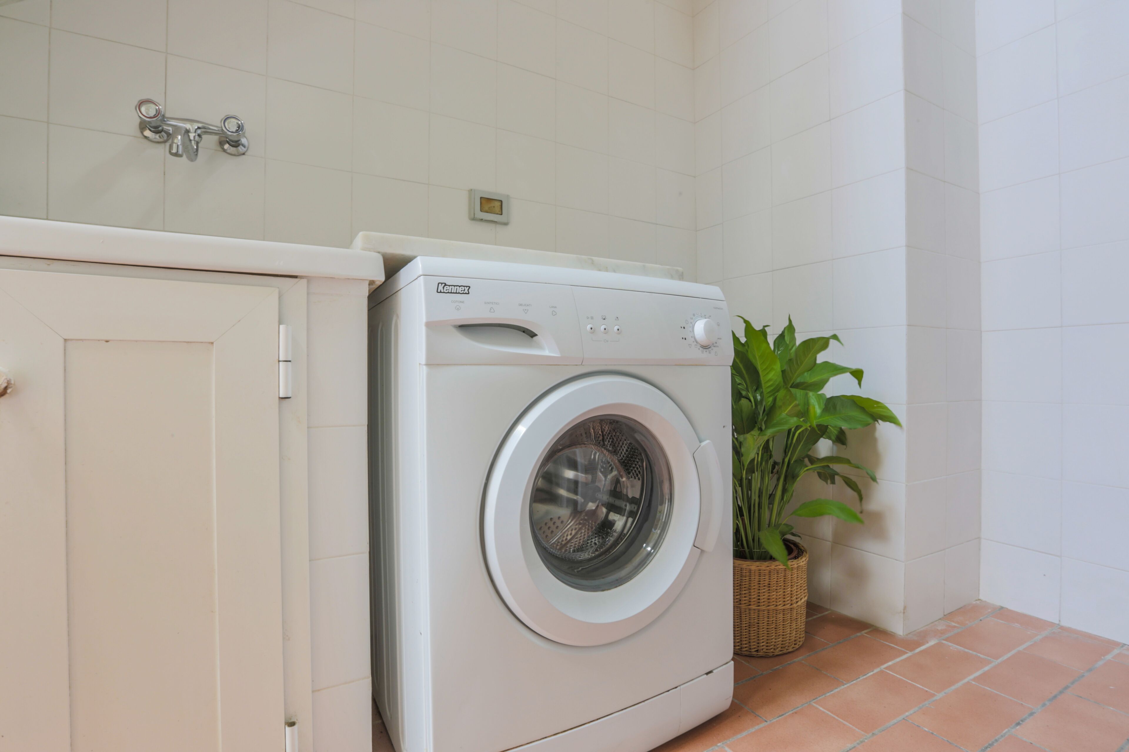 A compact, white washing machine in a small laundry area with a tiled wall and a potted plant.