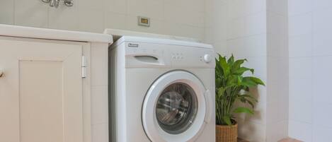 A compact, white washing machine in a small laundry area with a tiled wall and a potted plant.
