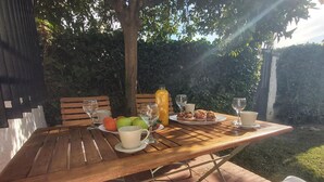 Ulkokäyttöön tarkoitettu puinen pöytä, jonka päällä on hedelmiä, muffinsseja ja juomia. Pöydän ympärillä on tuoleja ja penkki, ja taustalla näkyy puutarha ja puita.