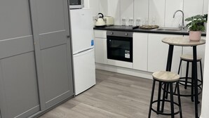 A modern kitchen with white cabinets, a grey refrigerator, a microwave, a stove, and a sink. There are two stools and a small table with a potted plant.