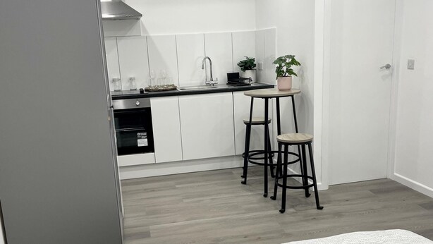 A compact bedroom with a bed, a small table with two chairs, and a kitchenette in the background.