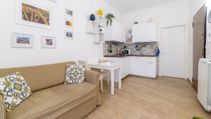 A modern living room with a beige sofa, a dining table, and a kitchen area with white cabinets and a tiled backsplash. - Cassari Cottage (Palermo)