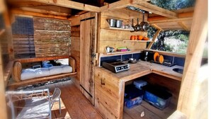 Une cabane en bois rustique, dotée d’une petite cuisine, d’un coin salon et d’une fenêtre donnant sur une zone boisée.