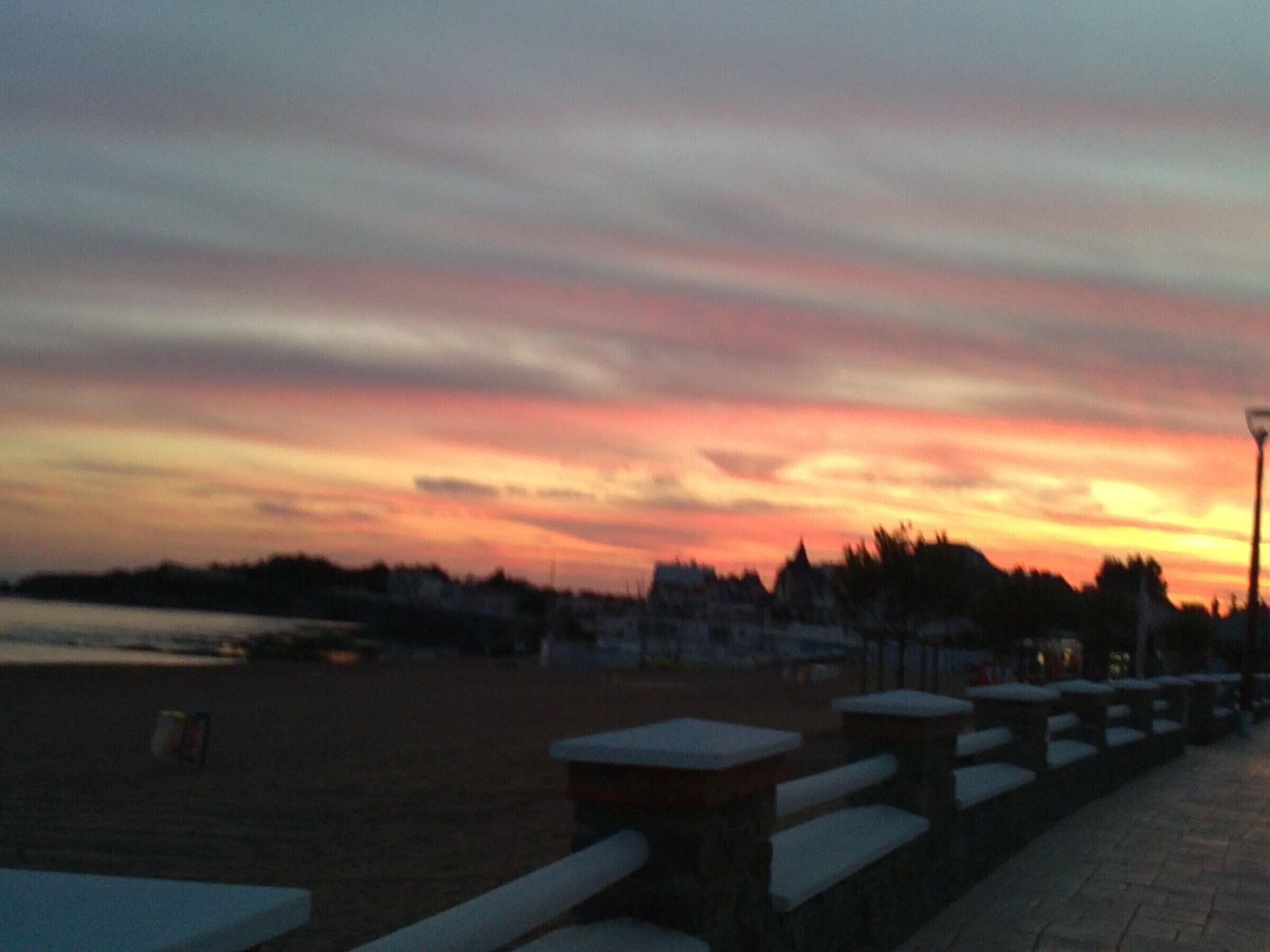 Solnedgang over en havnefront med bænke og en lygtepæl.