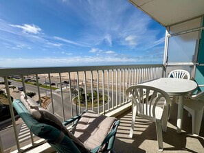 A balcony with a view of the beach, a table, and chairs.