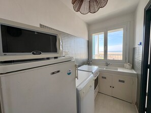 Eine kompakte Küche mit Mikrowelle, Kühlschrank und Waschmaschine. Ein Fenster bietet Blick auf den Strand.