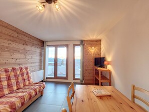 A cozy room with a wooden floor, a sofa, a wooden table with a book, a TV on a stand, and a door leading outside.