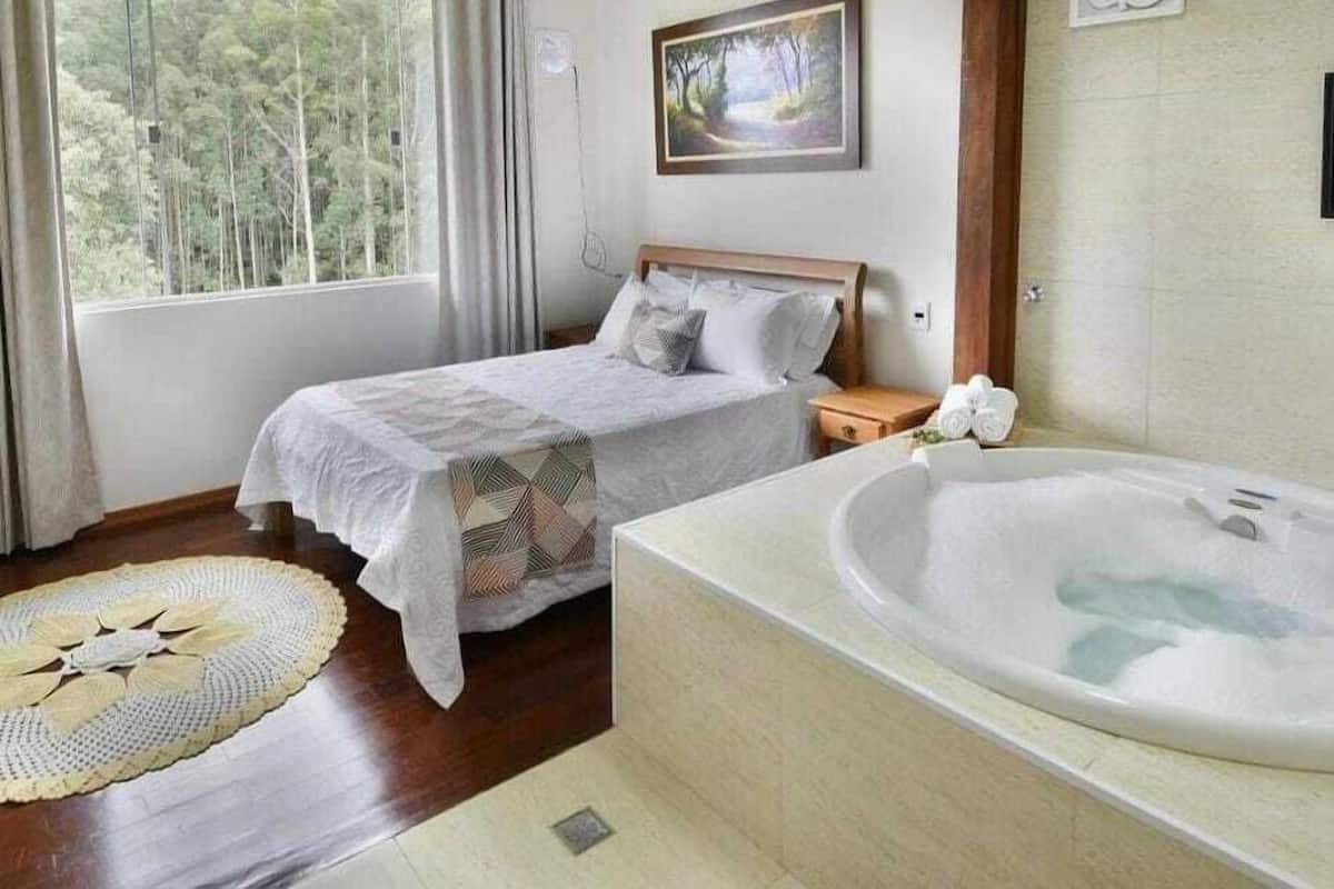 A bedroom with two beds, a large window, a painting, and a bathtub.