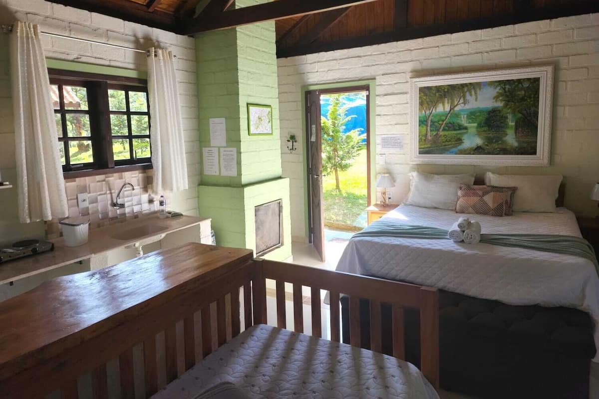 A room with a wooden bench, a bed with a green blanket, a painting on the wall, and a window with a view of greenery.