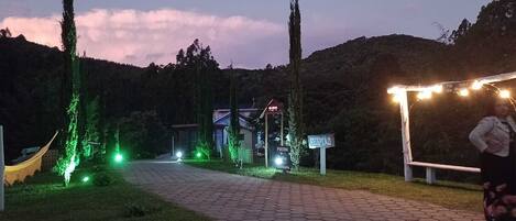 A paved pathway leading to a building with illuminated signage, surrounded by trees and a mountainous backdrop.