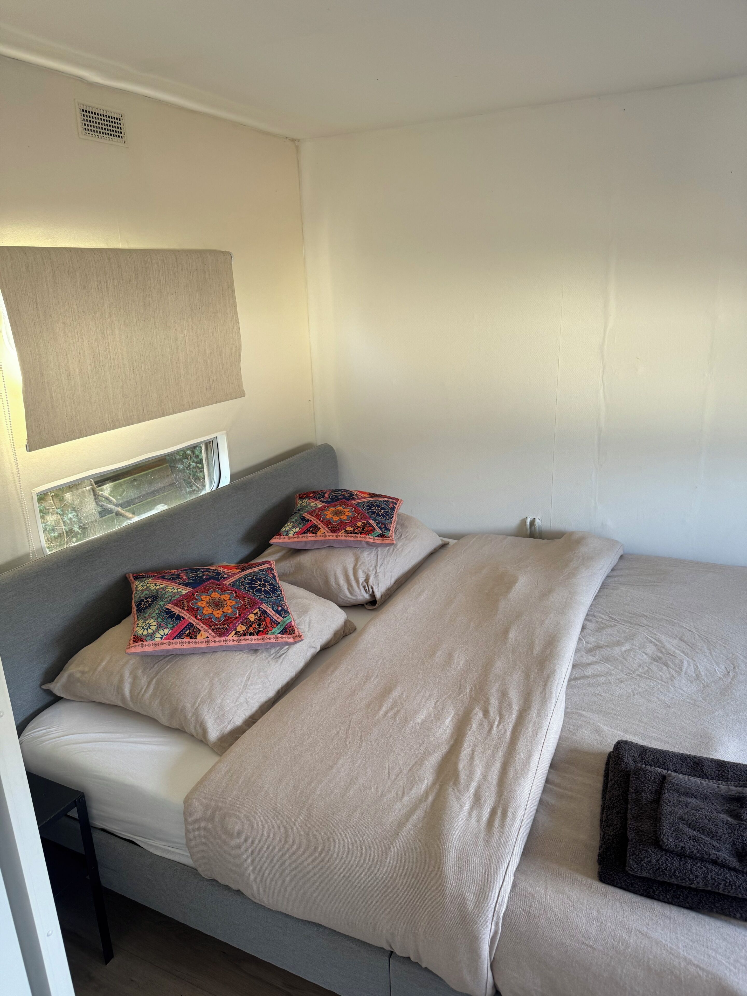A single bed with a grey headboard, two decorative pillows, a beige blanket, and a dark grey towel.