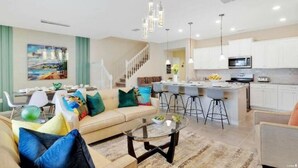 A modern living room with a sofa, coffee table, and a dining area with a bar. There is a kitchen in the background with white cabinets and a microwave. - 7495 OL - Solterra Resort (Davenport)