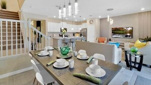 A modern kitchen with white cabinets, a dining area with a wooden table set for four, and a staircase with white railings. - 7495 OL - Solterra Resort (Davenport)