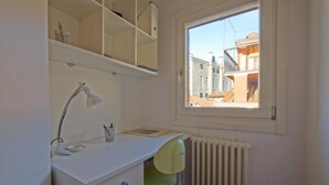 A small, bright room with a white desk, a green chair, a window with a view of buildings, and a radiator. - LA CASETTA (Venezia)