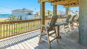 A wooden deck with two chairs and a table, overlooking a beach with houses and a flag. - Saltwater Soul - Cool Unique Mid-Level Hangout Area! (Surfside Beach)