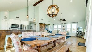 A spacious dining area with a wooden table and floral upholstered chairs, a kitchen in the background, and a ceiling fan. - Saltwater Soul - Cool Unique Mid-Level Hangout Area! (Surfside Beach)