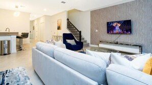 Un salon moderne avec un canapé, un fauteuil bleu, un téléviseur fixé au mur et un escalier en arrière-plan.