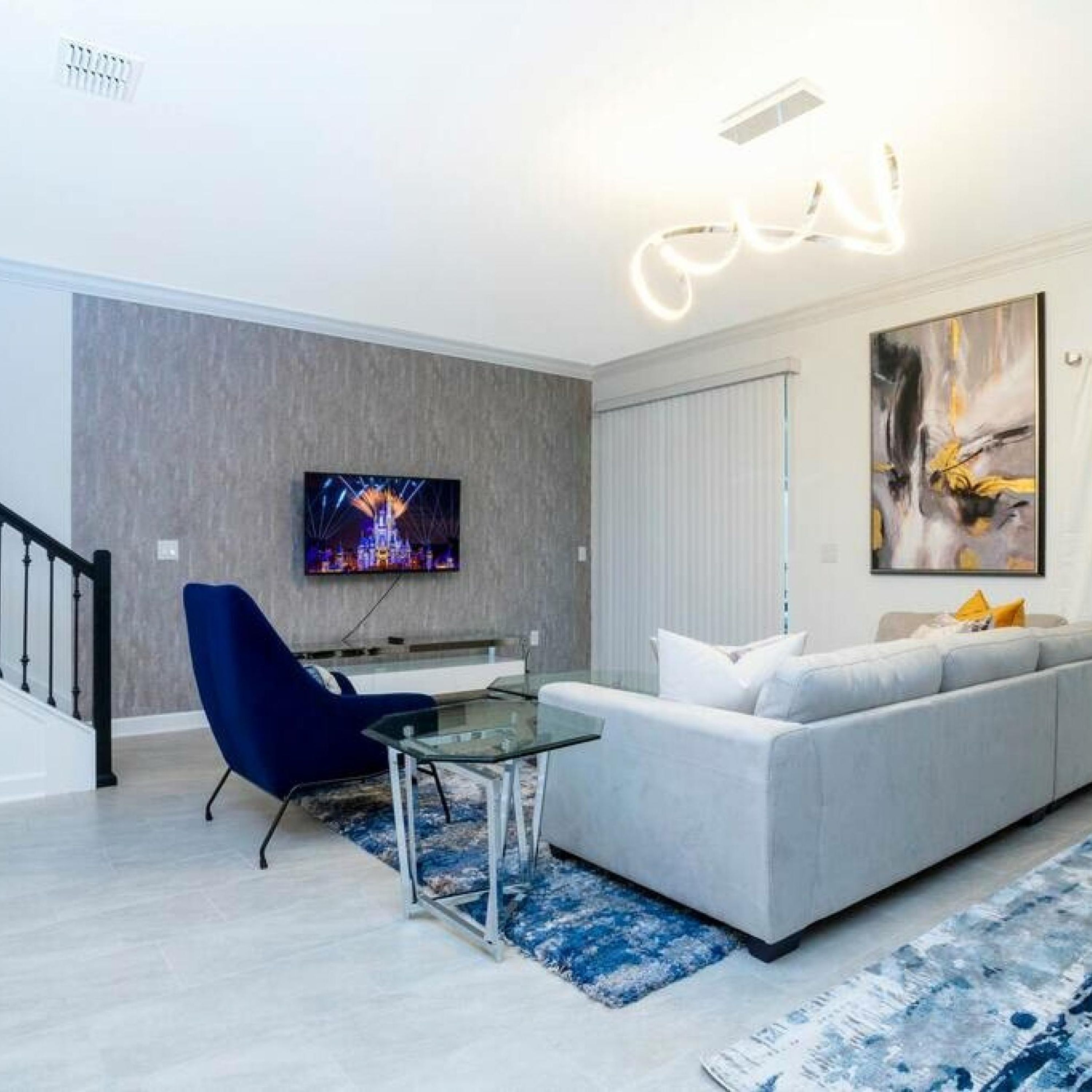 A modern living room with a grey sofa, a blue armchair, a glass coffee table, and a flat-screen TV mounted on the wall.