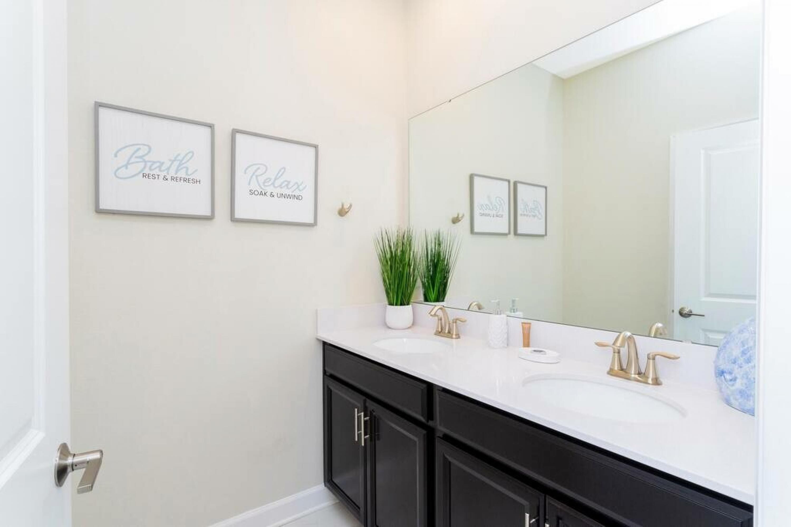 Une salle de bain avec un meuble-lavabo blanc, des armoires noires et un grand miroir. Deux panneaux encadrés sont accrochés au mur : l’un porte l’inscription « Espace détente et rafraîchissement » et l’autre « Savon et relaxation ».