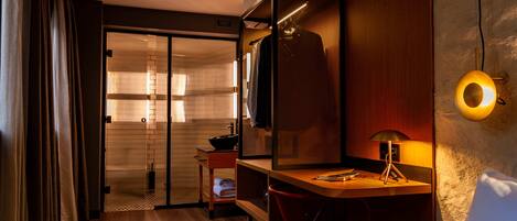A hotel room with a wooden desk, a red chair, a mirror, a small table with a lamp, and a sliding door leading to a bathroom.