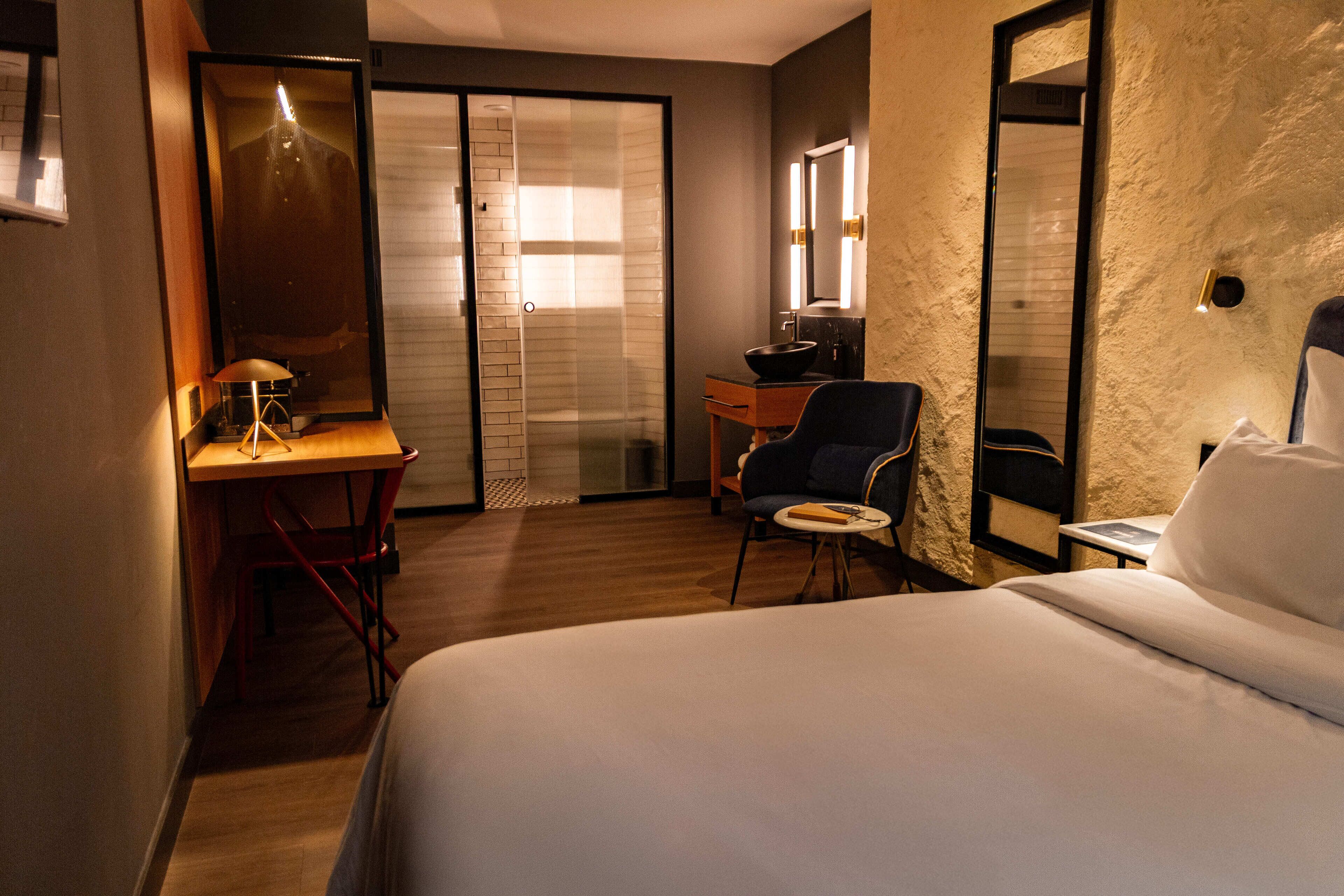 A hotel room with a bed, a desk, a chair, a small table, a mirror, and a sliding door leading to a shower area.