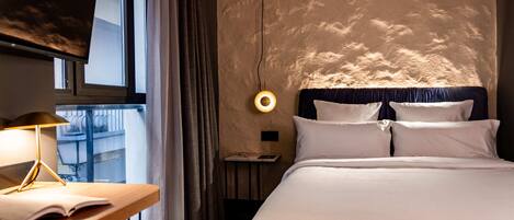 Quarto de hotel com uma cama, uma escrivaninha com luminária, uma janela com cortinas e um ponto de luz fixado na parede.