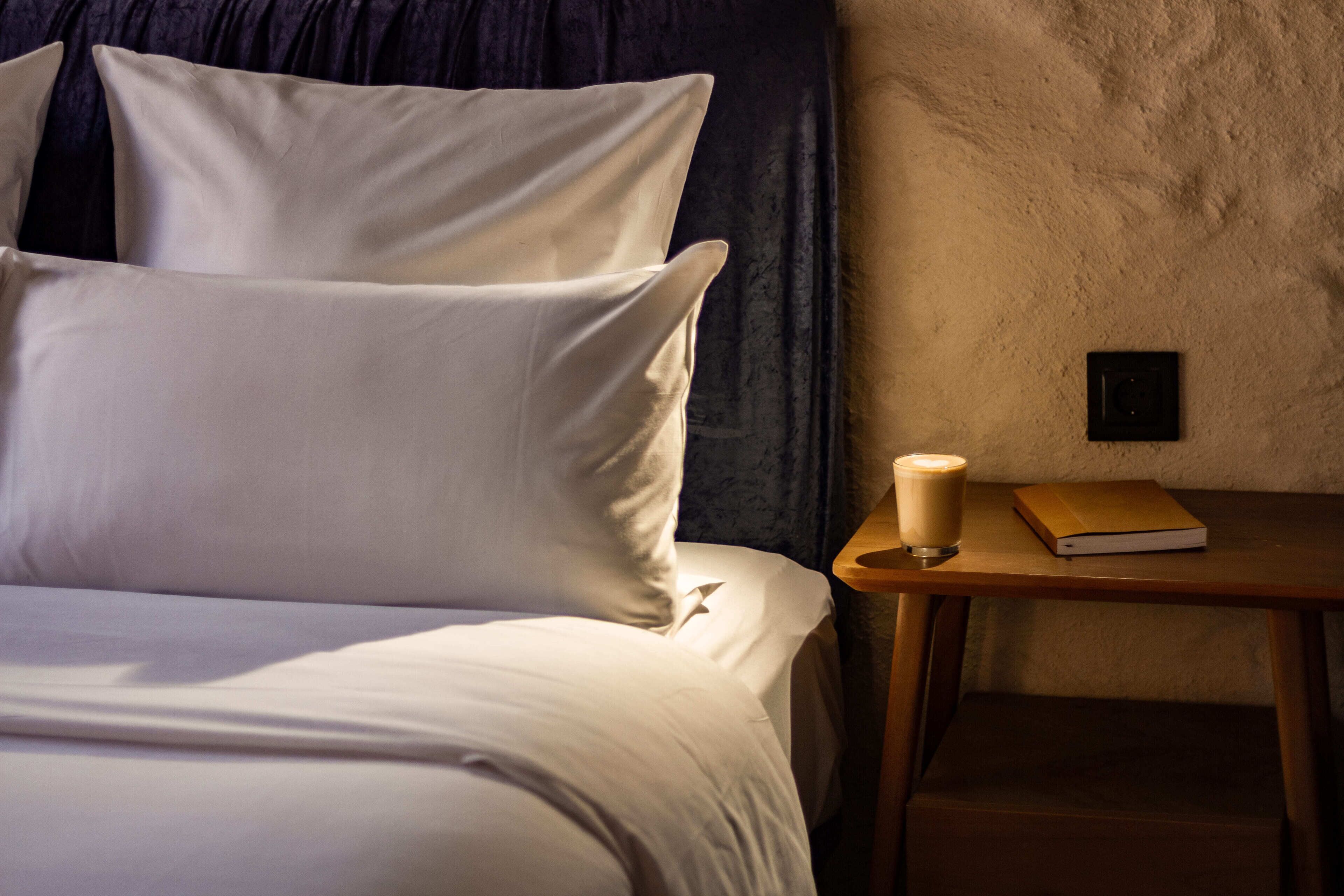 A neatly made bed with white linens and pillows, a bedside table with a book and a cup, and a textured wall in the background.