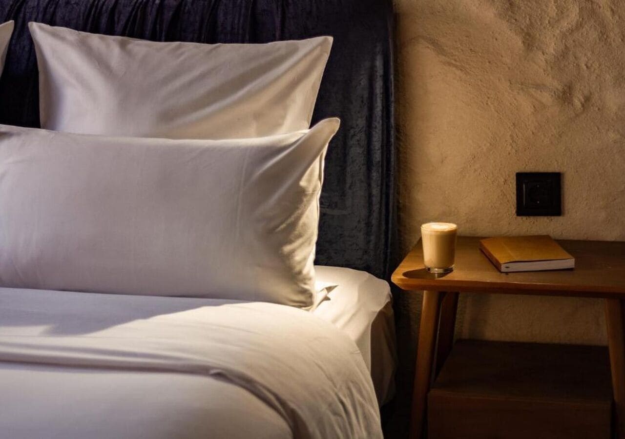 Un lit avec une literie blanche, une table de chevet avec un livre et une tasse, et un mur à texture.