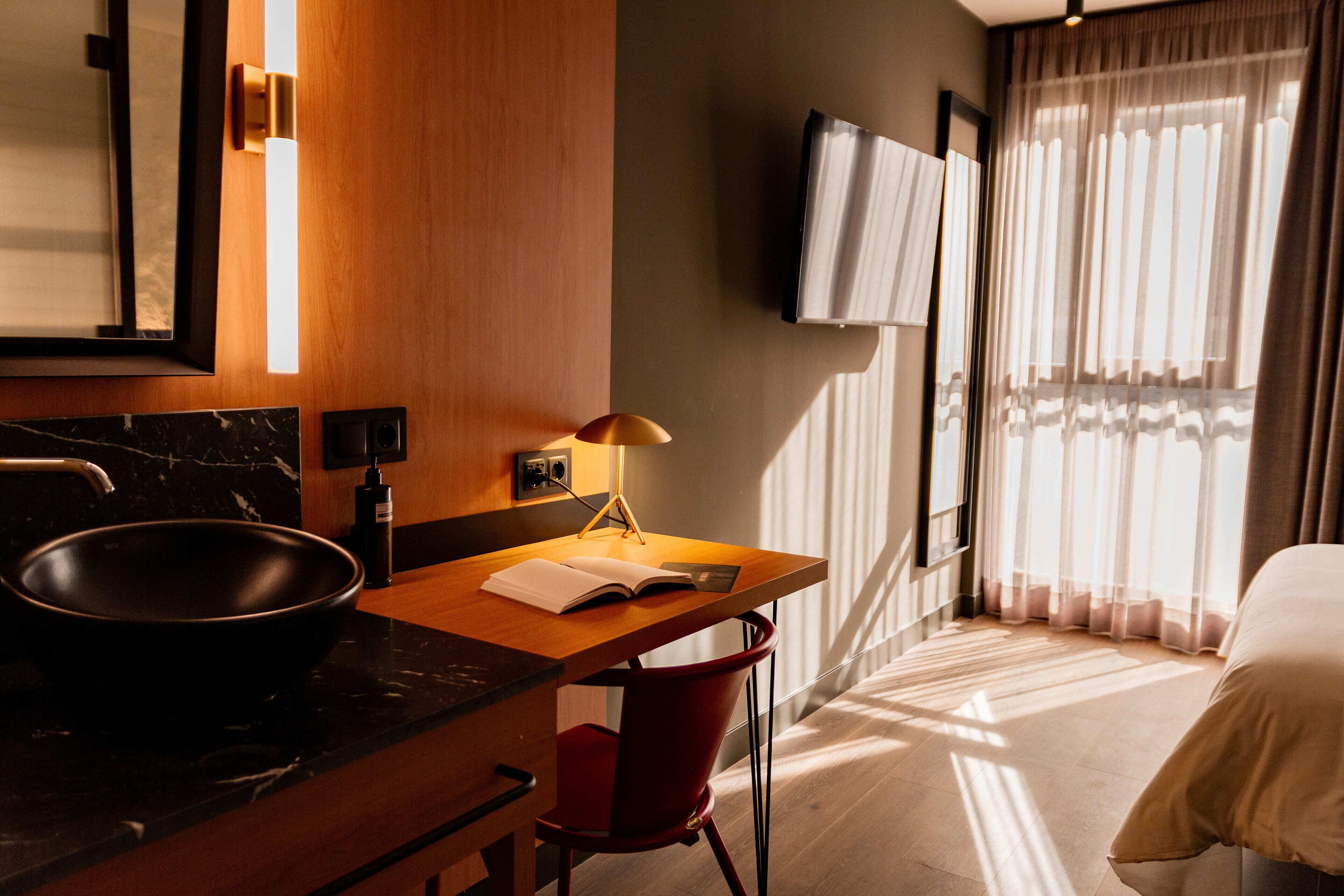 Una habitación de hotel moderna con un escritorio de madera, un lavabo negro, una silla roja, una cama y un televisor montado en la pared.