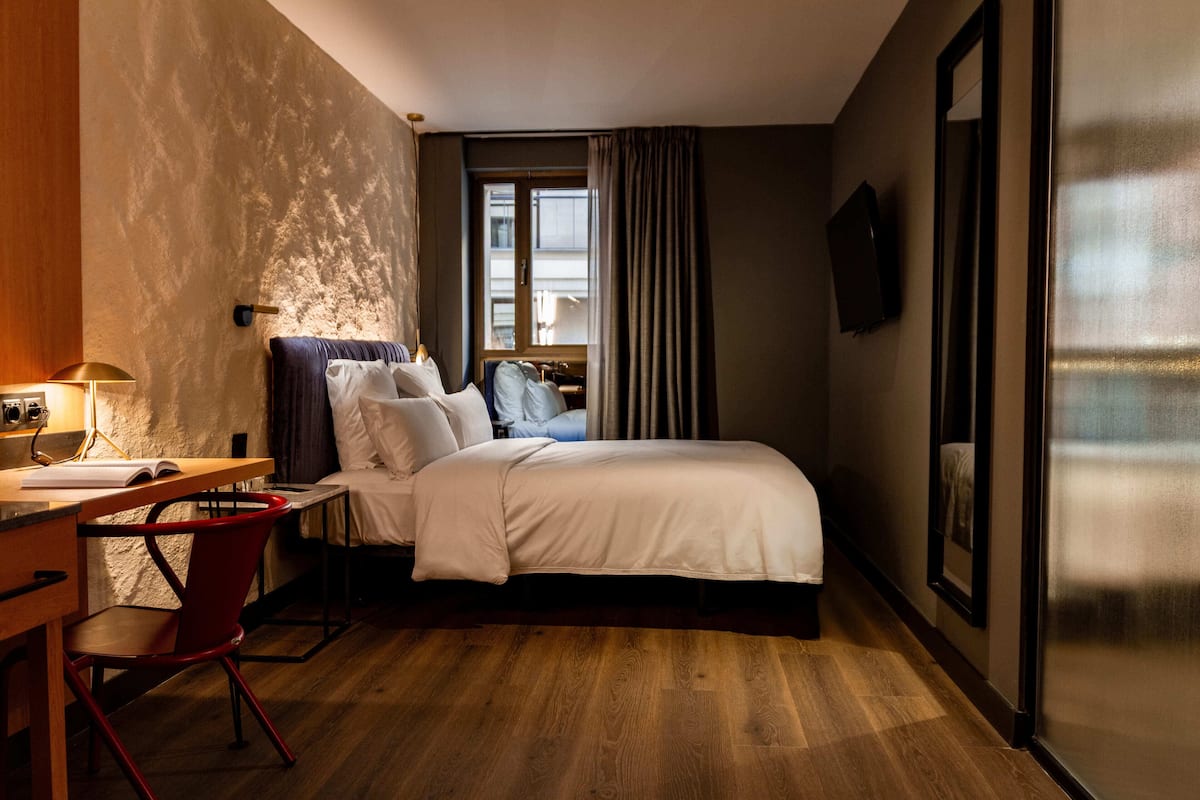 Une chambre d’hôtel comprenant un lit, un bureau avec une chaise, une fenêtre avec des rideaux et un téléviseur à écran plat fixé au mur.