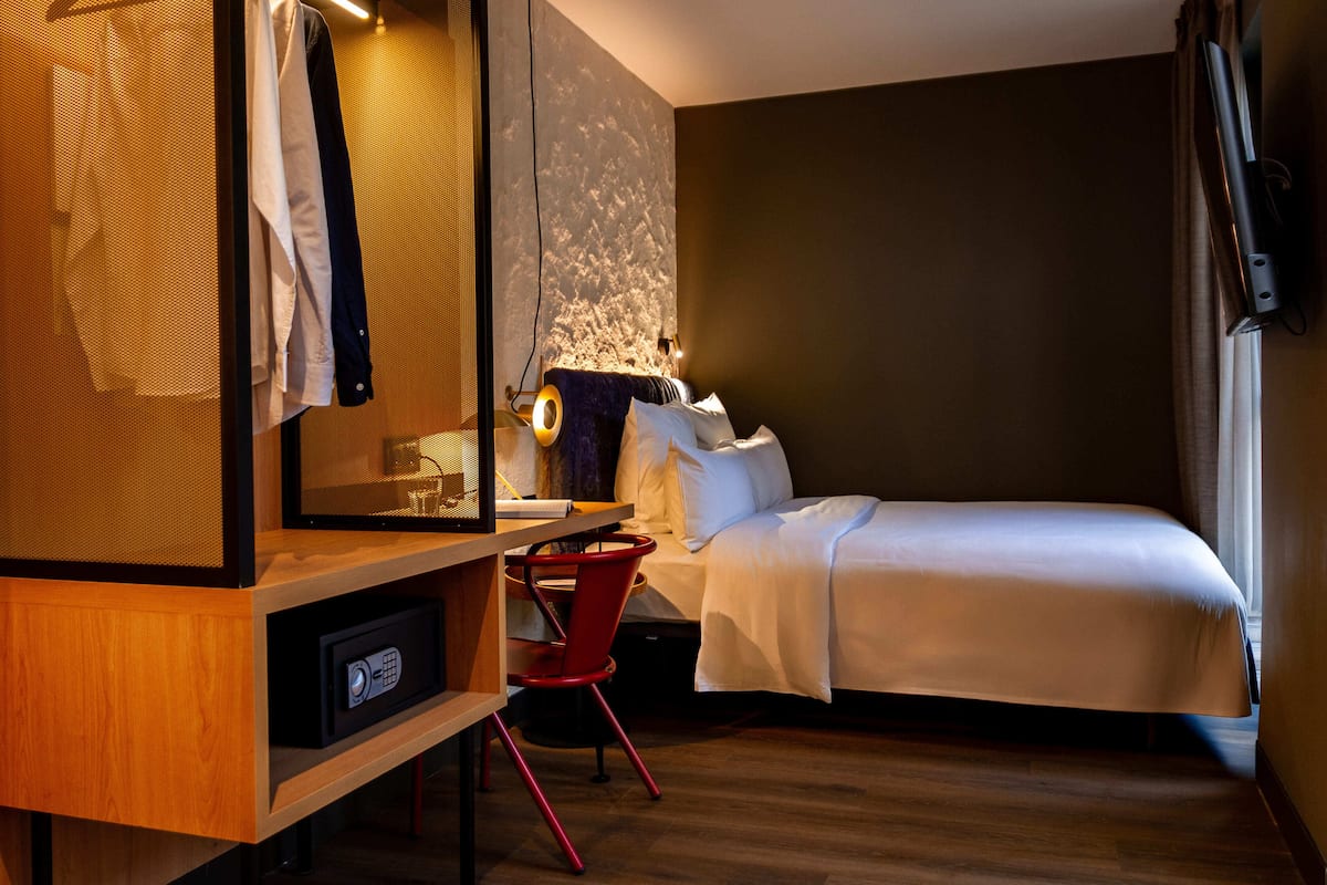 Une chambre d’hôtel comprenant un lit, un bureau avec une chaise, un téléviseur fixé au mur et une fenêtre avec des rideaux.
