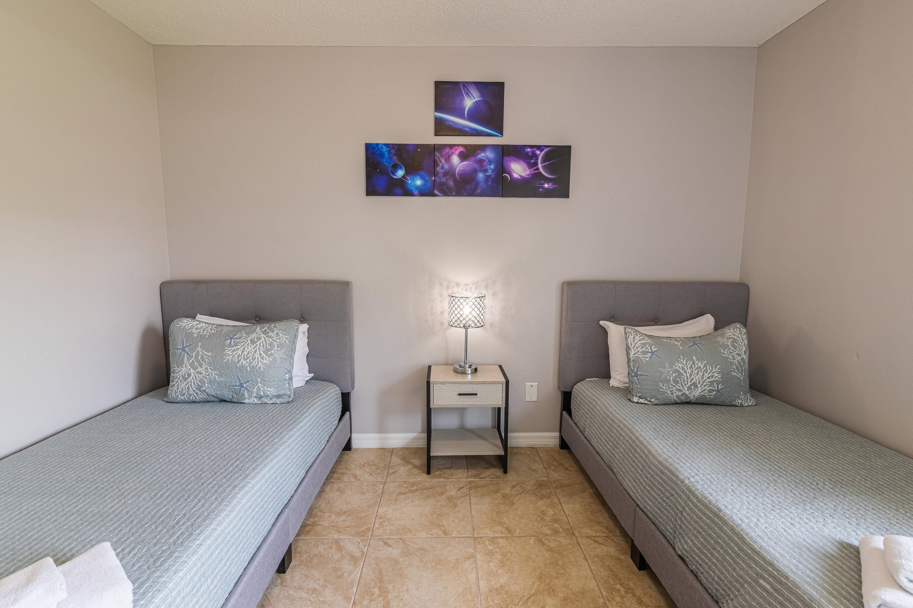 Een kleine, keurig opgemaakte slaapkamer met twee eenpersoonsbedden, een nachtkastje met een lamp en een kunstwerk aan de muur boven de bedden.