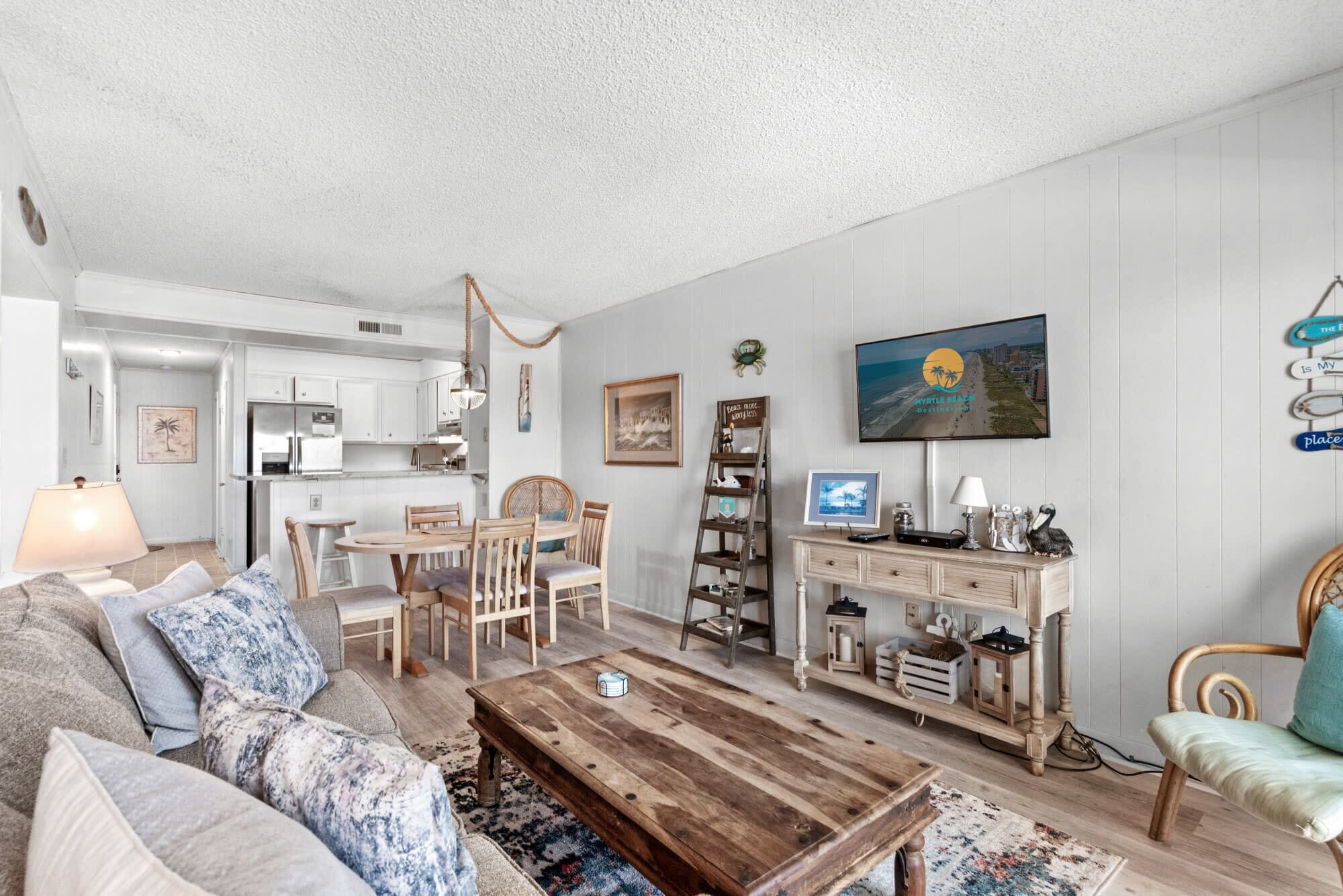 A living room with a sofa, coffee table, and dining area. There is a TV mounted on the wall, a ladder, and a desk with a computer.