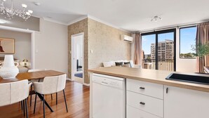 A modern kitchen with a dishwasher, a dining area with a wooden table and chairs, and large windows offering a view of the city. - Stunning 3BR Perth Penthouse (East Perth)