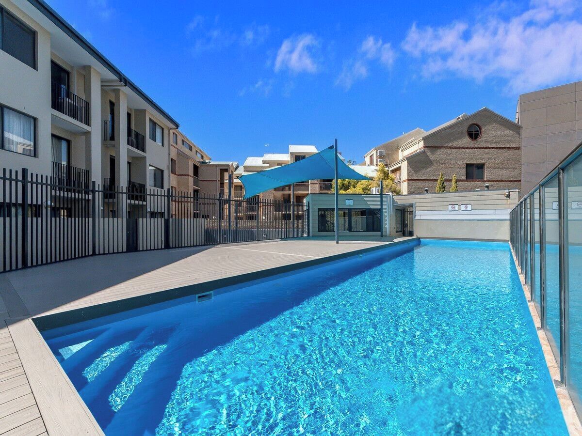 A swimming pool with a tiled deck, surrounded by buildings with balconies and a fence.