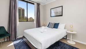 A modern hotel room with a large bed, a round side table, a green chair, and a view of the city through the window. - Stunning 3BR Perth Penthouse (East Perth)