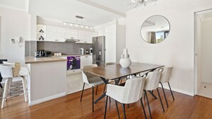 A modern kitchen with white cabinets, a central island, and a dining area with a wooden table and chairs. - Stunning 3BR Perth Penthouse (East Perth)
