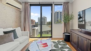 A modern living room with a white sofa, a glass coffee table, a TV, and a balcony with a view of the city. - Stunning 3BR Perth Penthouse (East Perth)