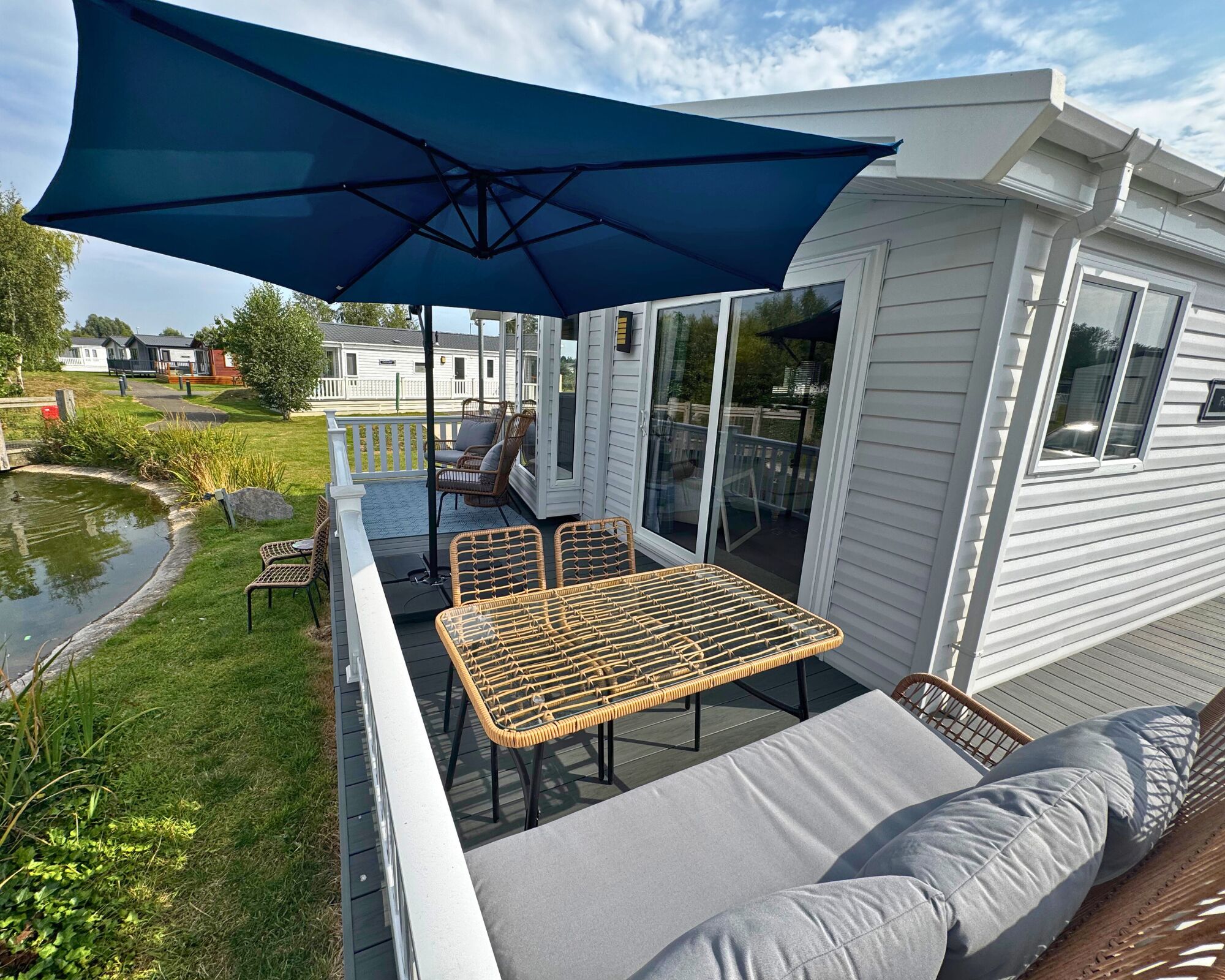 A covered outdoor deck with wicker furniture and a large umbrella, overlooking a pond and a residential area.