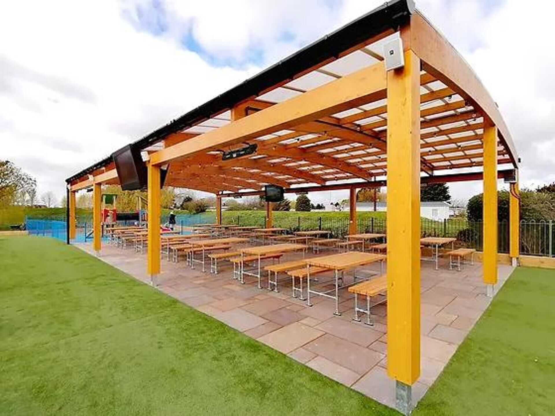 Un espace extérieur abrité, doté de tables et de bancs, entouré d’une pelouse verdoyante et avec une étendue d’eau en arrière-plan.