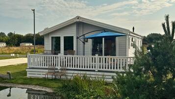 A single-story mobile home with a covered porch, a small deck, and a pond in the foreground.