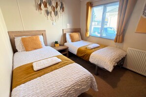 A hotel room with two single beds, each with a wooden headboard, white textured bedding, and a brown throw blanket. There is a small table with a plant between the beds, and a window with curtains to the right. - Sunset 19 - Southview Holiday Park Skegness (Skegness)