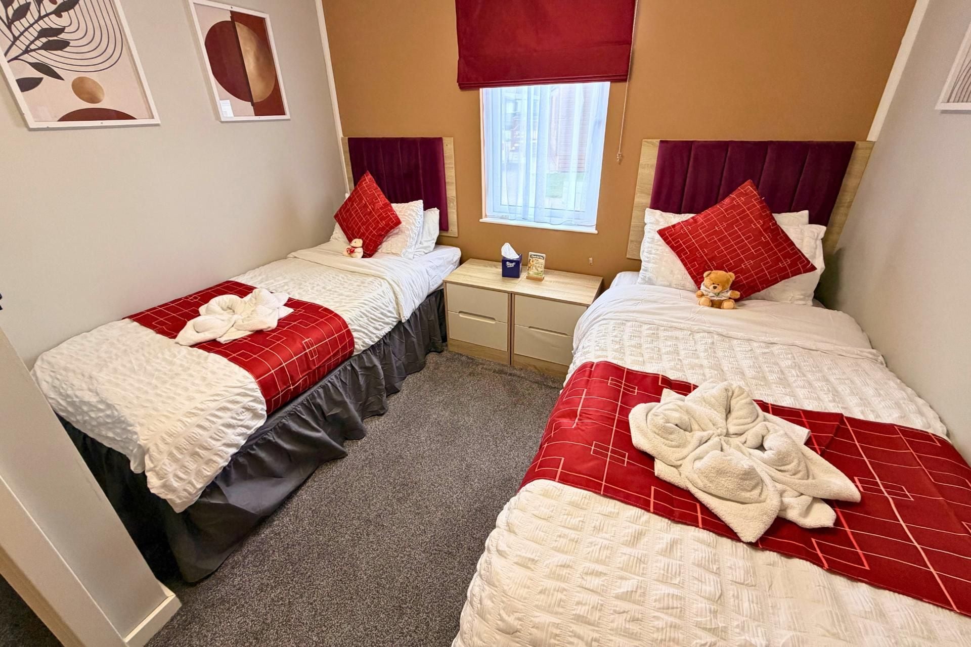 A hotel room with two beds, each with a red and white bedspread, a small bedside table with a tissue box, and a window with a red blind.