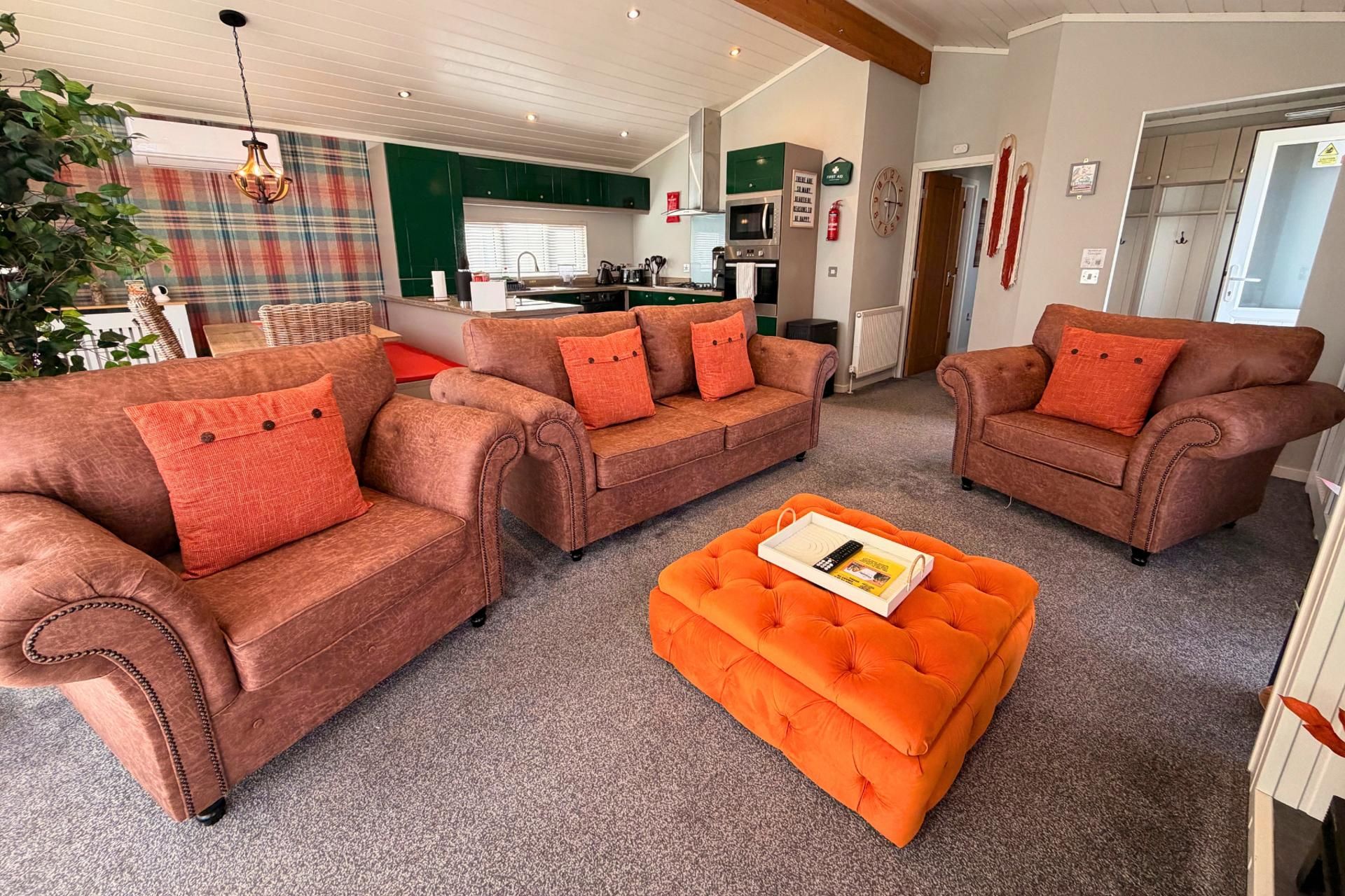 En stue med tre brune sofaer og en orange skammel, et køkken i baggrunden og en ternet gardin.