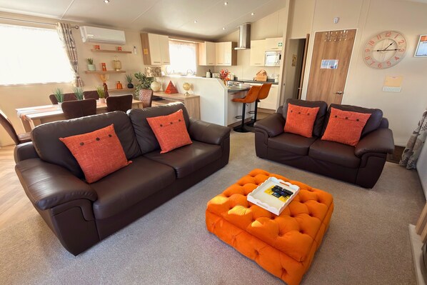 A modern living room with two dark brown sofas, each with red cushions, and an orange ottoman with a tray. - Sunset 19 - Southview Holiday Park Skegness (Skegness)