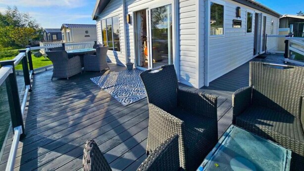A covered outdoor deck with wicker furniture and a glass table, adjacent to a white building with large windows and a covered entrance. - Sunset 19 - Southview Holiday Park Skegness (Skegness)