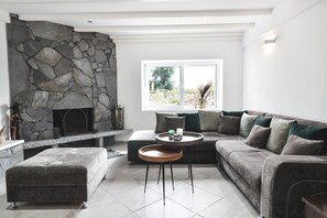A modern living room with a stone fireplace, a grey sofa, a round coffee table, and a large window with a view of greenery. - Moute House by Corfuescapes (Corfu)