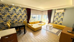 A living room with a yellow sofa, a dining table with chairs, and patterned wallpaper.